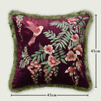 Botanical velvet cushion cover displayed upright, highlighting a detailed embroidered hummingbird and floral pattern in pink, coral, and green shades. The edges feature lush green fringe trim, making it a premium decorative pillow for sofas and accent chairs.