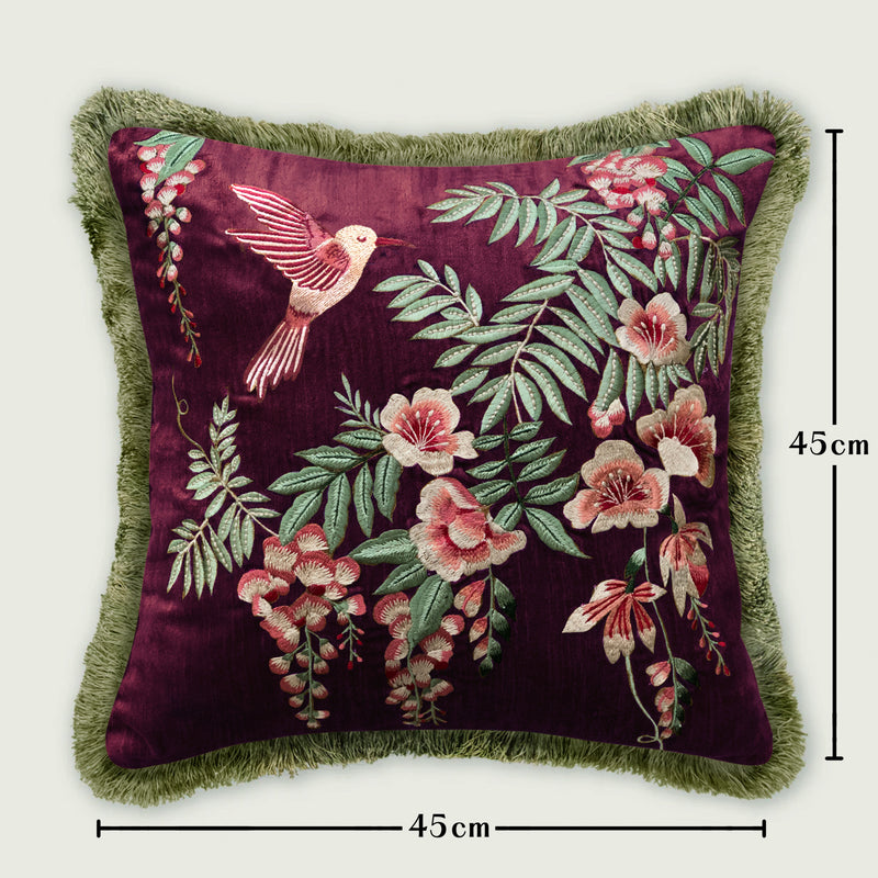 Botanical velvet cushion cover displayed upright, highlighting a detailed embroidered hummingbird and floral pattern in pink, coral, and green shades. The edges feature lush green fringe trim, making it a premium decorative pillow for sofas and accent chairs.