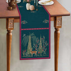 Whispering Grove blue table runner featuring intricate botanical artwork, a graceful deer motif and contrasting berry-pink borders, styled on a wooden dining table.