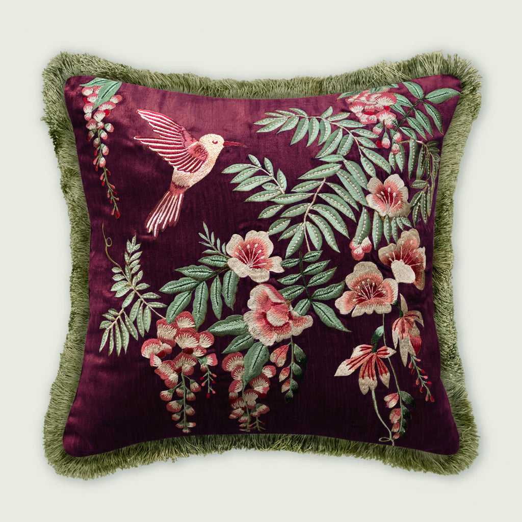 Wine velvet cushion featuring an embroidered hummingbird surrounded by detailed botanical florals and green foliage, finished with lush green fringe trimming. Designer decorative pillow ideal for sofas, beds, and home décor.